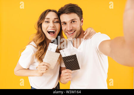 Portrait of happy couple man and woman in basic t-shirts holding travel tickets with passports while taking selfie photo isolated over yellow backgrou Stock Photo