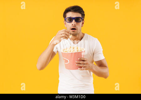 Photo of a concentrated young man in casual white t-shirt eat popcorn wearing 3d glasses watch film isolated over yellow background. Stock Photo