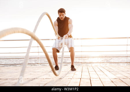 Photo of concentrated man in tracksuit working out with fitness rope on wooden pier at seaside in morning Stock Photo