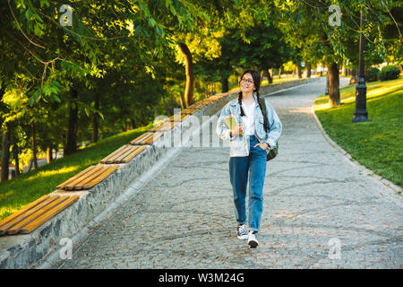 Happy young teenage student girl carrying backpack and books while walking outdoors at the park Stock Photo