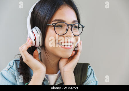 Portrait of a pleased happy pretty girl in denim jacket wearing eyeglasses isolated over gray background listening music with headphones. Stock Photo