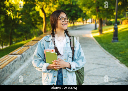 Happy young teenage student girl carrying backpack and books while walking outdoors at the park Stock Photo