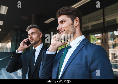 Image of confident businessmen partners dressed in formal suit standing outside office center and talking on cellphones during working meeting Stock Photo