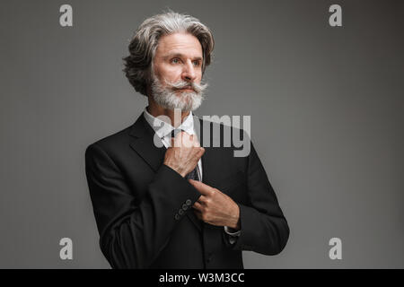 Image of handsome adult businessman wearing formal black suit looking aside while touching his tie isolated over gray background Stock Photo