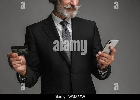 Cropped image of unshaven adult businessman wearing formal black suit holding credit card and smartphone isolated over gray background Stock Photo