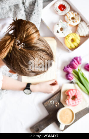 Girl reading a book and has brealfast in bed. Tray with strawberry pink donut with coffee and flowers. Background blurred. Top view. Stock Photo