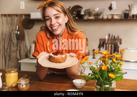 Picture of a happy cheery young blonde girl chef at the kitchen holding plate with sweet pastry. Stock Photo