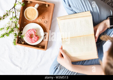 Spring brealfast in bed. Tray with strawberry pink donut with coffee and spring blossom. Girl reading a book. Top view. Copy space. Stock Photo