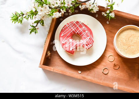 Spring brealfast in bed. Tray with strawberry pink donut with coffee and spring blossom. Girl reading a book. Top view. Copy space. Stock Photo