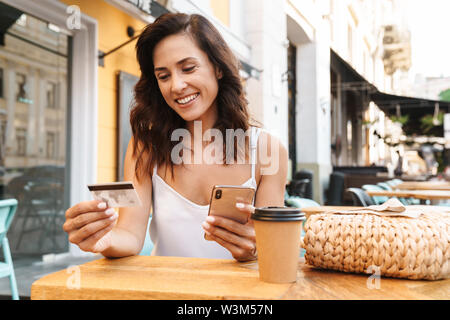 Portrait of smiling nice woman with straw bag holding credit card and using cellphone while sitting in cozy cafe outdoors Stock Photo