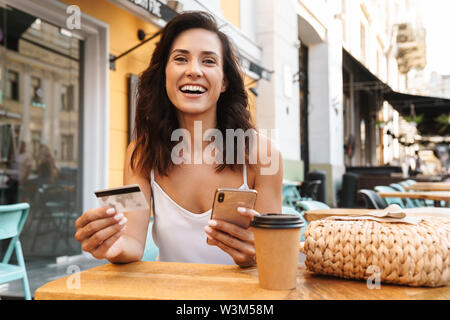 Portrait of delighted nice woman with straw bag holding credit card and using cellphone while sitting in cozy cafe outdoors Stock Photo