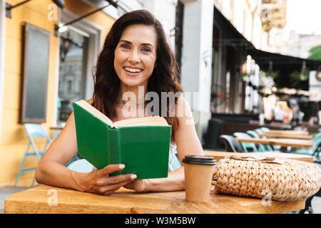 Portrait of positive nice woman with straw bag drinking coffee from paper cup and reading book while sitting in cozy cafe outdoors Stock Photo