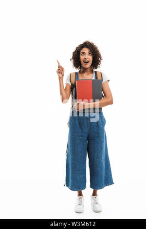 Image of surprised african american woman wearing backpack pointing finger at copyspace and holding books isolated over white background Stock Photo