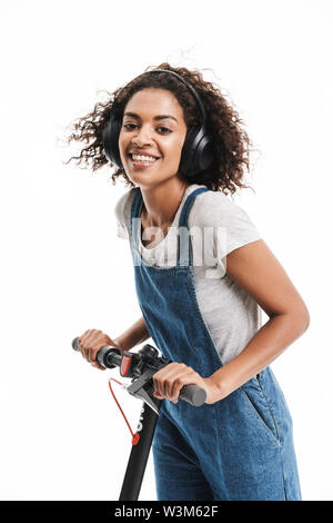 Image of pretty african american woman dressed in denim overalls using headphones while riding on scooter isolated over white background Stock Photo