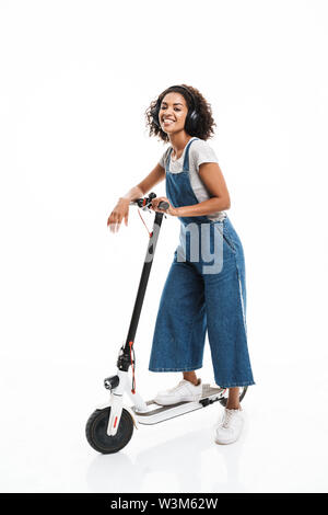 Image of smiling african american woman dressed in denim overalls using headphones while riding on scooter isolated over white background Stock Photo