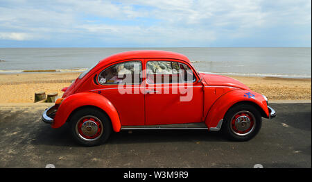 Classic Red  Volkswagen Beetle parked on seafront promenade with sea and beach in background. Stock Photo
