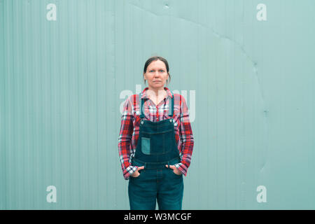 Serious female farmer posing on farm. Confident woman farm worker wearing plaid shirt and jeans overalls looking at camera, with copy space Stock Photo