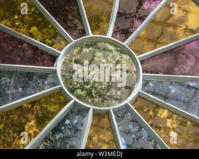 Coloured pink, yellow and blue glass creates an interesting effect on the foliage seen through the spokes of a wheel-shaped stained glass window. Stock Photo
