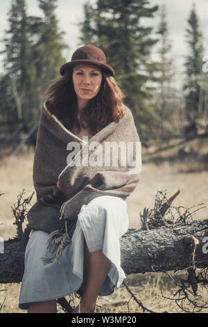 An attractive young woman sits on a log wrapped in a woollen blanket. Yukon Territory, Canada Stock Photo