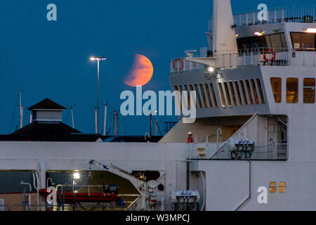 Southampton, Hampshire, England, UK, 16th July 2019. A partial lunar eclipse and a hazy sky on the horizon turn the moon orange red during evening moonrise over the south coast. In the foreground, the Red Falcon Red Funnel Isle of Wight ferry has just docked in the port for disembarkation. Credit: Paul Biggins/Alamy Live News