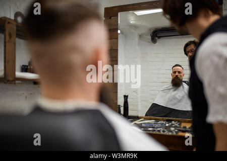 Brutal guy in modern Barber Shop. Hairdresser makes hairstyle a man with a long beard. Master hairdresser does hairstyle with hair clipper Stock Photo