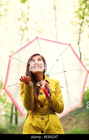 Asian Autumn woman happy after rain walking with umbrella. Female model looking up at clearing sky joyful on rainy fall day wearing yellow raincoat outside in nature forest. Multiracial Asian girl. Stock Photo