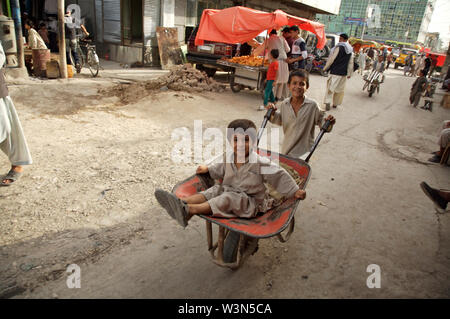 A young boy wheels his younger brother on a cart. They work as porter, at the main market, in the city of Mazar-e-Sharif, in the northern province of Balkh. An estimated 60,000 children have fled broken families, poverty and conflict, and now live or work in the streets of major cities. Afghanistan. June 26, 2007. Stock Photo