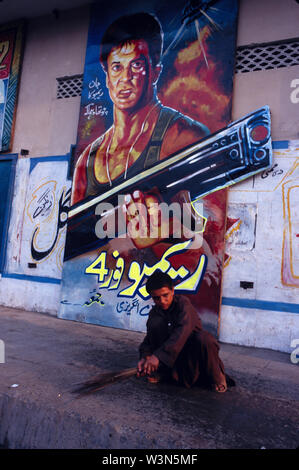 A young Afghan boy sweeps floor clean, in front of a larger than life size poster of the popular action movie Rambo, in Kissa Khuwani Bazaar, in Peshawar, the capital of North West Frontier Province, in Pakistan. Stock Photo