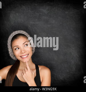Thinking woman by blackboard. Multiracial hipster university college student pensive and contemplating looking up to the at empty blank copy space on chalkboard black background. Asian Caucasian, 20s. Stock Photo