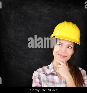 Woman architect, engineer, surveyor or construction worker wearing protection hardhat thinking looking thoughtfully to the side on chalkboard blackboard texture for copy space. Stock Photo