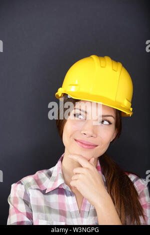 Young female architect, engineer, surveyor or construction worker wearing hardhat thinking as she standing with hand to chin looking thoughtfully to the side, studio portrait on a dark background Stock Photo