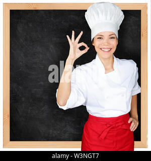 Chef showing menu blackboard and Perfect hand sign. Woman in front of blank menu blackboard. Happy female chef, cook or baker by empty chalkboard menu display wearing chef whites uniform and hat Stock Photo
