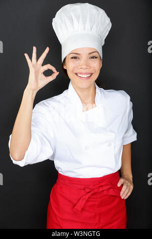 Chef woman giving a Perfect gesture with hand. Young beautiful female chef with a beaming smile standing in a toque chefs hat and whites and apron giving a Perfect gesture with her fingers ion black. Stock Photo