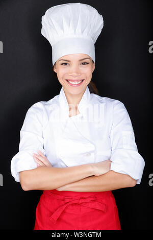 Asian female chef in chef whites uniform and hat. Woman chef, cook or baker portrait on black background. Young multiracial Chinese Asian / Caucasian female model standing proud and cross-armed. Stock Photo