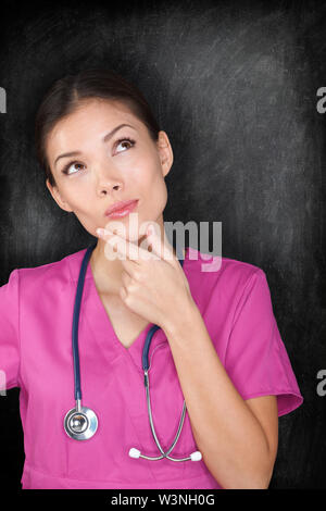 Female medical doctor nurse thinking by blackboard. Woman medical professional looking thinking at empty chalkboard copy space. Multiracial Asian Caucasian nurse in pink scrubs. Stock Photo