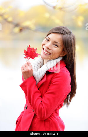 Fall girl holding red Autumn leave outside. Asian woman outdoor portrait in red seasonal autumn coat by fall forest lake. Female model smiling happy looking at camera. Mixed race Asian Caucasian girl. Stock Photo