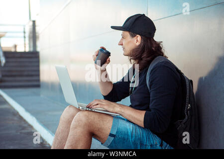 Young man with long hair and baseball cap sits on yellow skateboard on the asphalt, leaning his back on gray granite wall, and and drinks coffee. Stock Photo