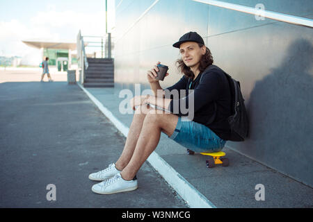 A young man in a baseball cap, with a backpack, and in denim shorts, drinks coffee while sitting on a yellow skateboard on the asphalt. Stock Photo