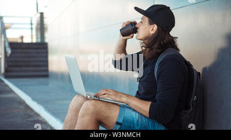 Young man with long hair and baseball cap sits on yellow skateboard on the asphalt, leaning his back on gray granite wall, and and drinks coffee. Stock Photo