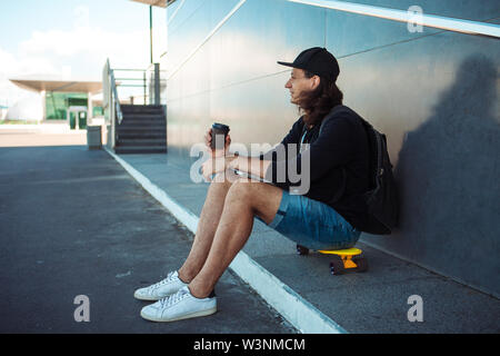 A young man in a baseball cap, with a backpack, and in denim shorts, drinks coffee while sitting on a yellow skateboard on the asphalt. Stock Photo