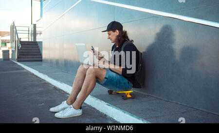 A young man, sitting on a yellow skateboard, is trying to synchronize a laptop with a phone. Apparently it happened. He happily look into the frame. Stock Photo