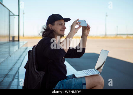 A young brunette man with long hair sits on the granite steps in the shade, with a laptop on his lap and makes a photo on a smartphone. Stock Photo