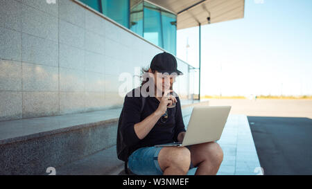 A young brunette man with long hair is sitting on the granite steps with a laptop spread out on his lap and looking thoughtfully into the distance. Stock Photo