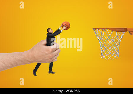 Big male hand holding businessman with basketball ball reaching out to hoop on yellow background Stock Photo