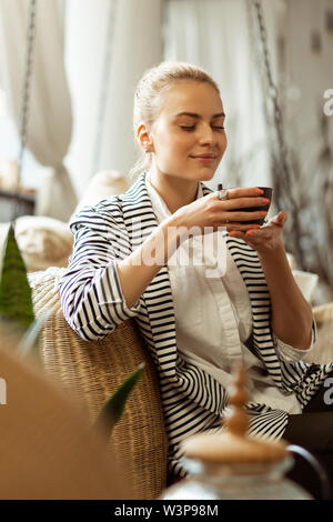 Chilling in living room. Coquettish blonde woman in elegant outfit resting on a couch with fresh green tea Stock Photo