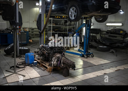 An sedan car raised on a lift for repair and under it a detached engine suspended on a blue crane and a gear box on a lifting table in a vehicle repai Stock Photo