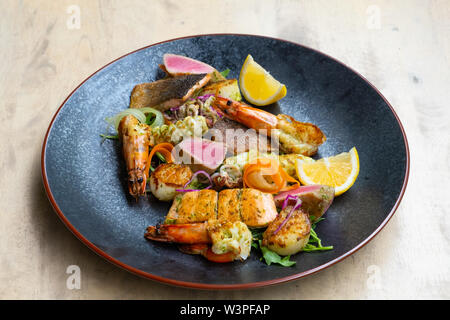 close up of mix grilled seafood and fish platter with fresh salad, prawns, scallop, tuna, salmon, seabass, lemon, green sauce in a black plate profess Stock Photo