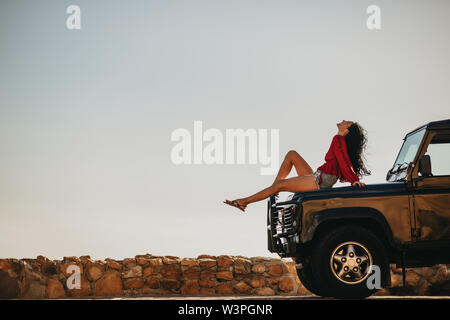 Side view of young woman sitting on car hood and looking up. Female taking a break on the road trip. Stock Photo