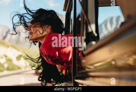 Beautiful woman sitting on car with her face out of the window with her hair flying in air. Female enjoying the car ride on a road trip. Stock Photo