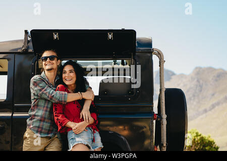Couple standing beside their car on a highway. Man embracing his woman while standing outdoors and looking away smiling. Stock Photo